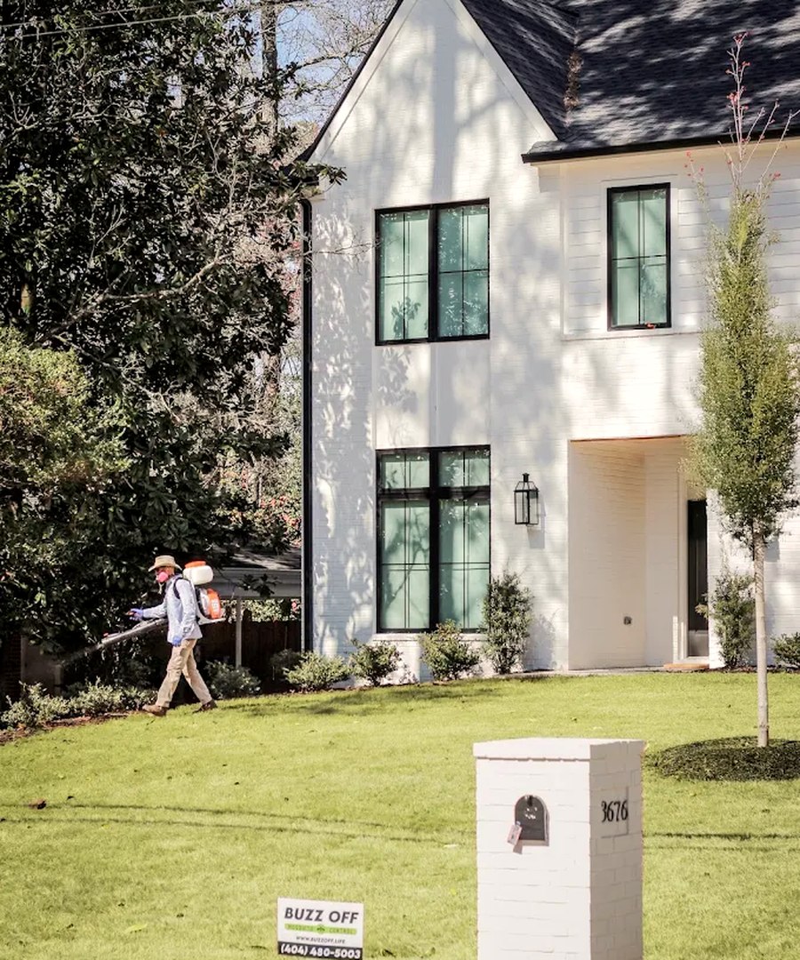 A Buzz Off technician treating the yard of a modern white Atlanta home, with a Buzz Off sign out front
